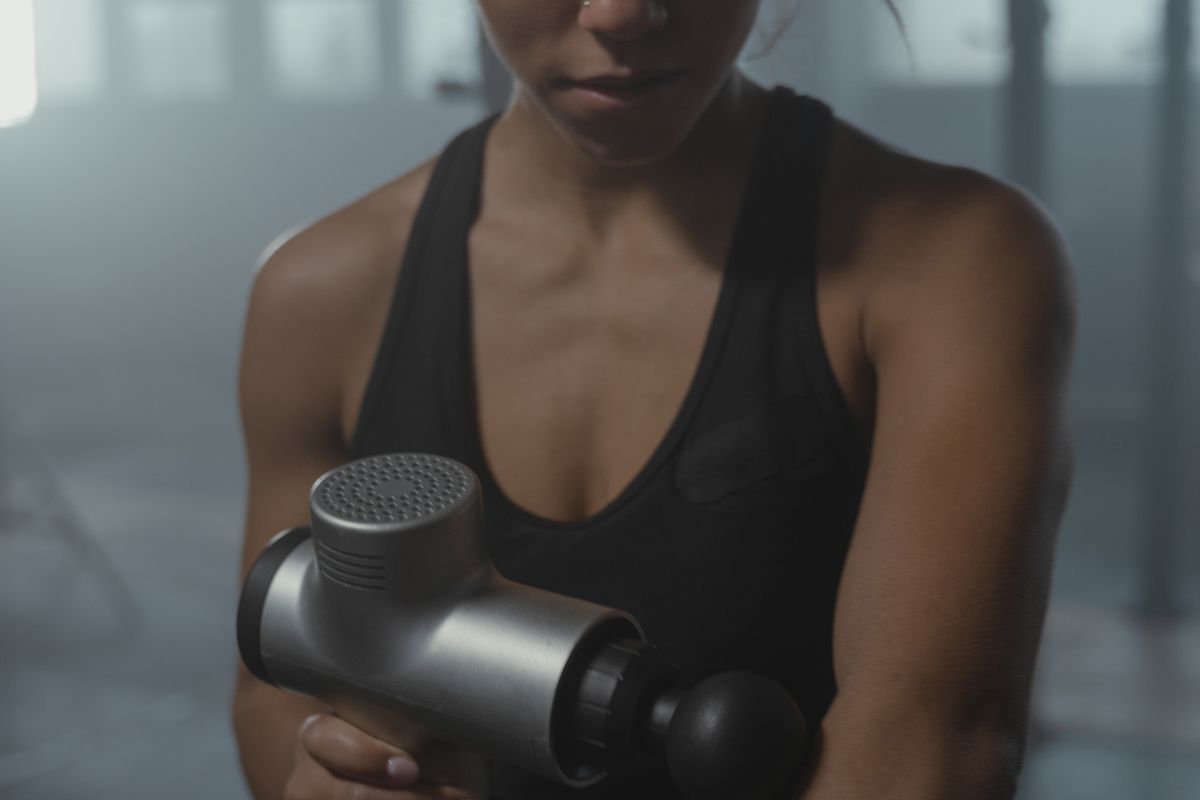 A person holding a massage gun against their forearm in a gym